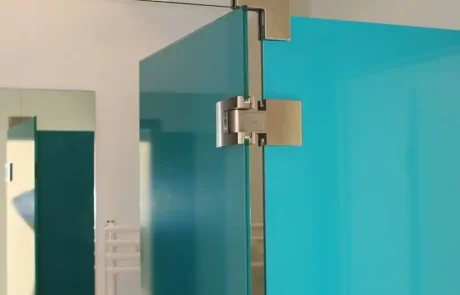 glaserei-wolff-duschabtrennung Nahaufnahme einer gläsernen Duschtür mit Edelstahlscharnieren, einer türkisfarbenen Glasscheibe und einem Teil eines Badezimmers im Hintergrund.