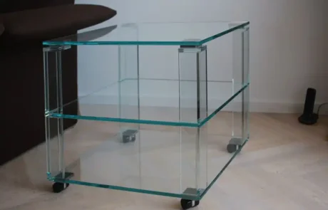 glaserei-wolff-glastisch Ein Beistelltisch aus Klarglas mit zwei Ablagen und vier Rollen, der auf einem hellen Holzboden neben einem dunkelbraunen Sofa steht.