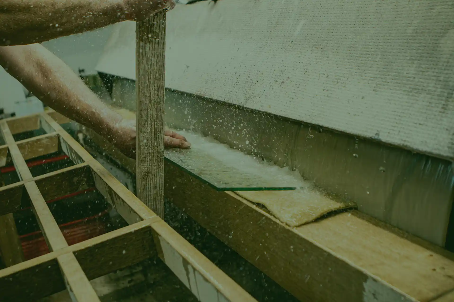 Eine Person stellt eine Glasscheibe auf einem Holzrahmen unter einen Wasserstrahl, wobei das Wasser auf der Oberfläche spritzt.