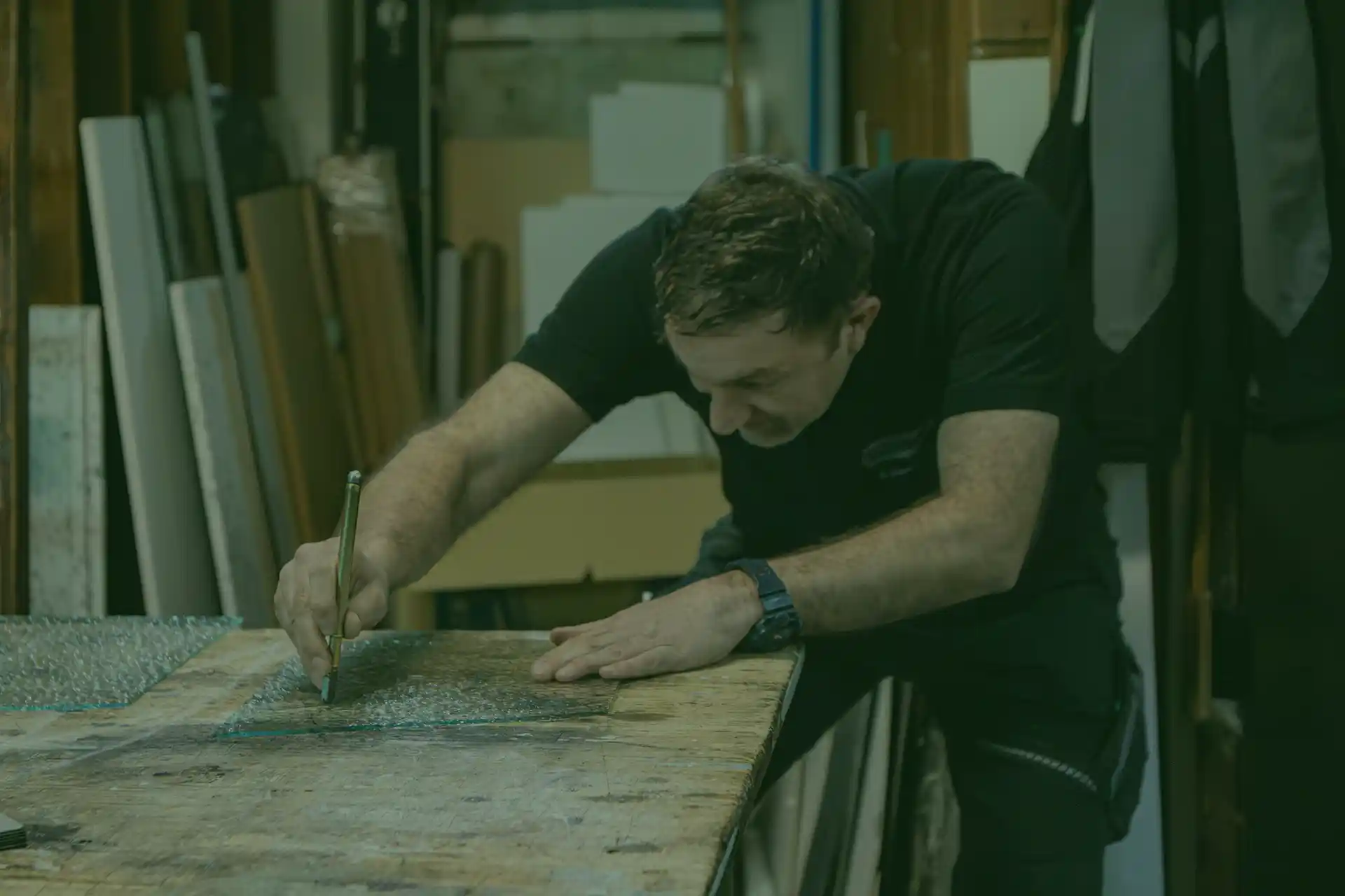 Ein Mann beugt sich über eine Werkbank und schneidet eine Glasscheibe mit einem Werkzeug in einer Werkstatt voller Werkzeuge und Materialien.